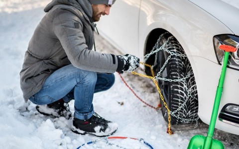 Mann mit Mütze und dicker Jacke kniet im Schnee und zieht am Auto vorne rechts Schneeketten auf
