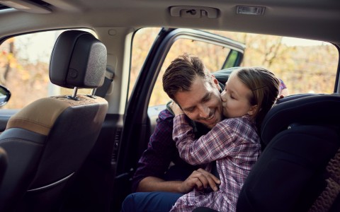 Vater setzt seine kleine Tochter in den Kindersitz auf der Rückbank eines Autos, während diese ihm einen Kuss auf die Wange gibt, beide tragen karierte Hemden