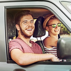 Zwei junge Männer sitzen lachend im Auto, der Fahrer trägt einen Strohhut und Sonnenbrille, der Beifahrer hat den Arm auf das geöffnete Fenster gelegt