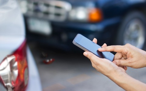 Männliche junge Hände tippen auf einem Handy, im Hintergrund sieht man unscharf zwei Fahrzeuge, die kollidiert sind