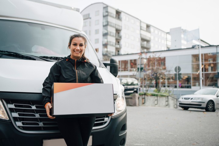 Eine lächelnde Frau lehnt sich an einen Lieferwagen und hält ein Paket