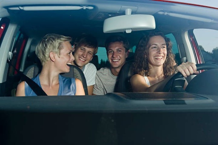 Vier junge Leute - zwei Frauen vorne, zwei Männer hinten - lachen alle fröhlich zusammen, während sie im Auto ungterwegs sind