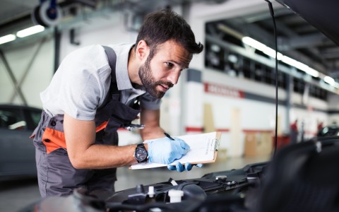 Mechaniker mit grauer Latzhose beugt sdich in den Motorraum eines Autos, das in einer Werkstatt steht und inspiziert etwas