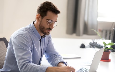 Mann mit Bart und hellblauem Hemd sitzt am Schreibtisch mit einem Laptop und schreibt etwas auf einen gelben Zettel