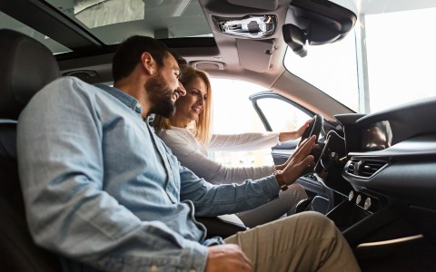 Junge blonde Frau sitzt am Steuer eines stehenden Autos und schaut auf die Armaturen, während ein Mann mit Dreitagebart ihr dort lächelnd etwas erklärt
