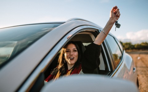 Junge dunkelhaarige Frau lacht und reckt den Arm mit Schlüssel in der Hand triumphierend aus dem geöffneten Fahrerfenster