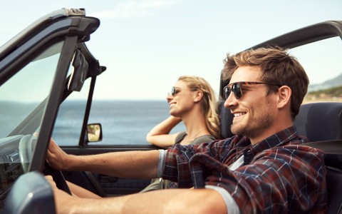 Päärchen fährt glücklich lächelnd in einem Cabrio bei Sonnenschein am Meer entlang, das im Hintergrund zu sehen ist