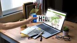Männliche Hand hält ein Kennzeichen mit H-DI in die Kamera, im Hintergrund ein Laptop, auf dem die Kfz-Versicherungsseite von HDI zu sehen ist