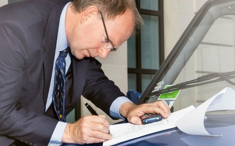 Mann im Anzug mit Brille unterschreibt ein Schriftstück auf der Motorhaube eines Autos