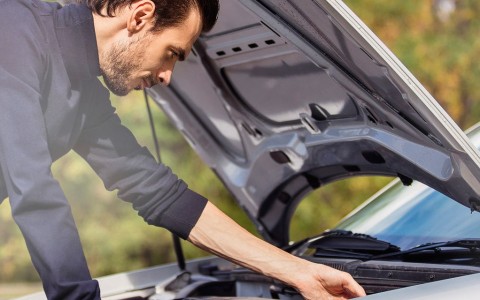 Mann in Jeans stützt sich bei geöffneter Motorhaube auf seinem Auto ab und überprüft etwas im Motorraum