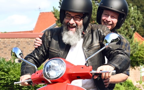 Mann mit langem grauen Bart, schwarzer BikerLederjacke und Helm sitzt lachend  auf einer roten Vespa mit einer Frau mit Helm hinter sich, die ebenfall lacht.