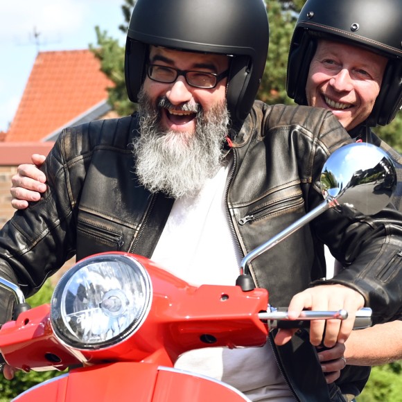 Mann mit langem grauen Bart, schwarzer BikerLederjacke und Helm sitzt lachend  auf einer roten Vespa mit einer Frau mit Helm hinter sich, die ebenfall lacht.