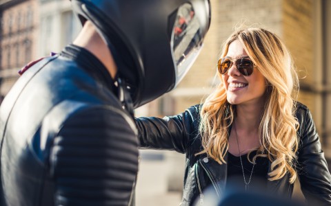 Blonde Frau mit langen Haaren, Sonnenbrille und schwarzer Lederjacke unterhält sich lachend mit einem männlichen Motorradfahrer, der einen schwarzen Helm trägt