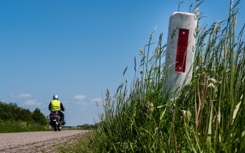 Motorradfahrer von hinten mit neongelber Warnweste fährt auf einem älteren Motorrad über eine Asphaltstraße, die beideseitig von Gras gesäumt ist, die Sonne scheint