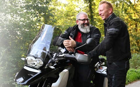 Zwei Männer in schwarzer Motorradkleidung, einer mit langem grauen Bart sitzt auf seinem abgestellten Motorrad, während ein jüngerer daneben steht