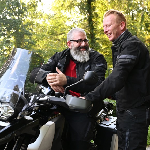 Zwei Männer in schwarzer Motorradkleidung, einer mit langem grauen Bart sitzt auf seinem abgestellten Motorrad, während ein jüngerer daneben steht