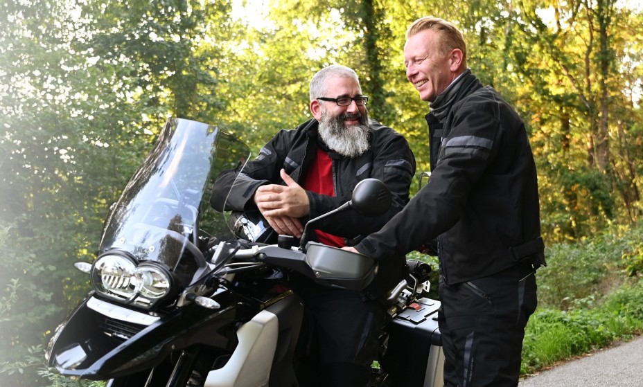 Zwei Männer in schwarzer Motorradkleidung, einer mit langem grauen Bart sitzt auf seinem abgestellten Motorrad, während ein jüngerer daneben steht