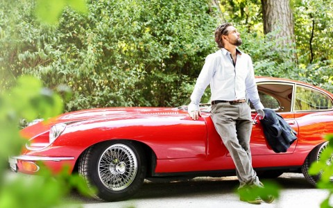 Mann lehnt entspannt an seinem knallroten Sport-Oldtimer, der am Wegrand im Wald parkt