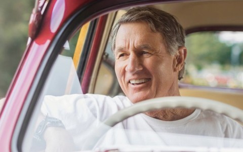 Älterer Mann in einem roten Oldtimer mit dem Lenkrad auf der rechten Seite, hat den Arm lässig aus dem Fenster gelehnt und lächelt in den Außenspiegel