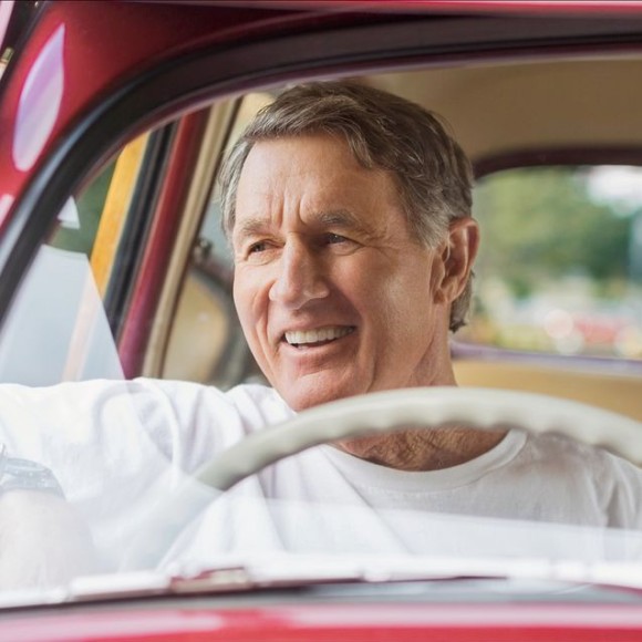 Älterer Mann in einem roten Oldtimer mit dem Lenkrad auf der rechten Seite, hat den Arm lässig aus dem Fenster gelehnt und lächelt in den Außenspiegel