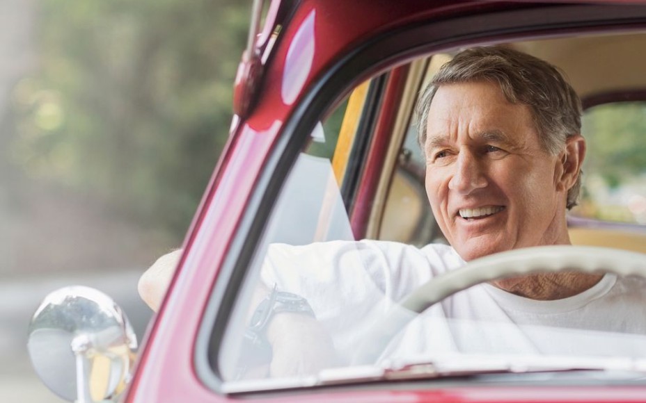 Älterer Mann in einem roten Oldtimer mit dem Lenkrad auf der rechten Seite, hat den Arm lässig aus dem Fenster gelehnt und lächelt in den Außenspiegel