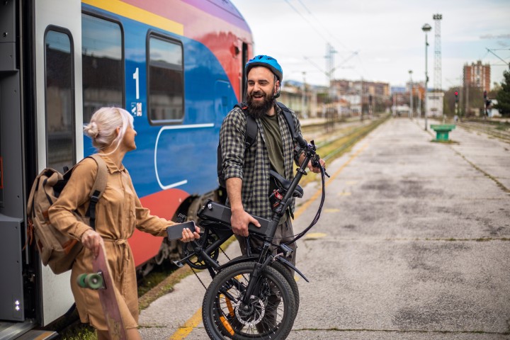 Mann mit Faltrad und blauem Helm steht lachend neben einer Frau mit Skateboard und beide sind gerade aus einerm Zug gestiegen, der noch hinter ihnen steht.