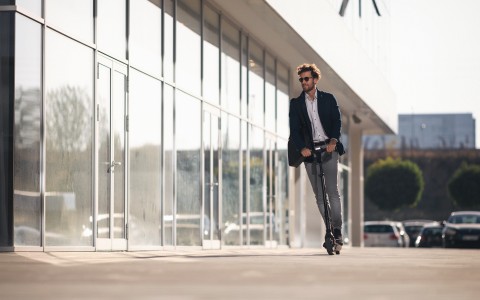 Mann mit Sonnenbrille fährt auf einem E-Roller an einer großen Fensterfront von Büros vorbei
