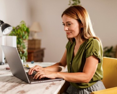 Junge Frau auf gelbem Polsterstuhl sitzt an einem Tisch an ihrem Laptop und tippt etwas