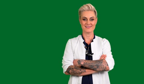 Doc Caro steht mit verschränkten Armen in weißem Arztkittel vor grünem Hintergrund