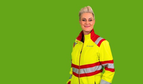 Doc Caro steht in neongelber Rettungssanitäterjacke vor hellgrünem Hintergrund und läßt die Arme locker an der Seite herunterhängen