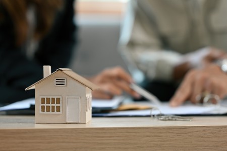Kleines Model-Haus aus Holz steht auf einer hellbraunen Holz-Tischplatte, im Hintergrund unschaf sind zwei Mneschen zu sehen, die Zettel und Stifte haben und etwas kalkulieren