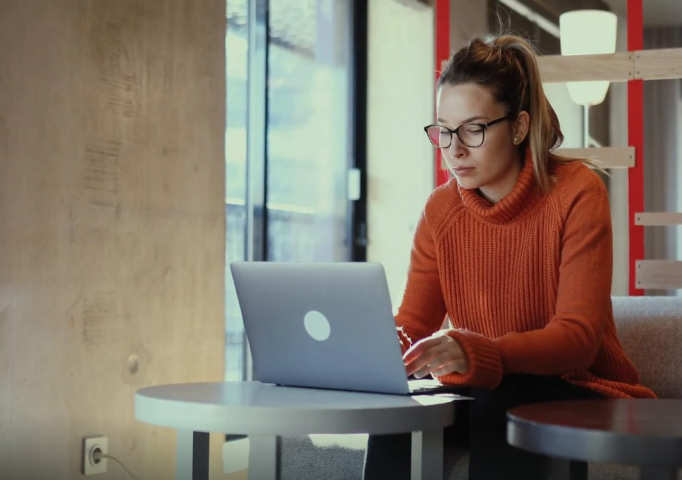 Blonde Frau mit Pferdeschwanz, Brille und orangefarbenem Pullover sitzt an einem Beistelltisch und wendet sich wahrscheinlich ans Beschwerdemanagement einer Versicherung. Im Hinterrgrund moderner Raum mit Loft-Charakter und roten Akzenten.
