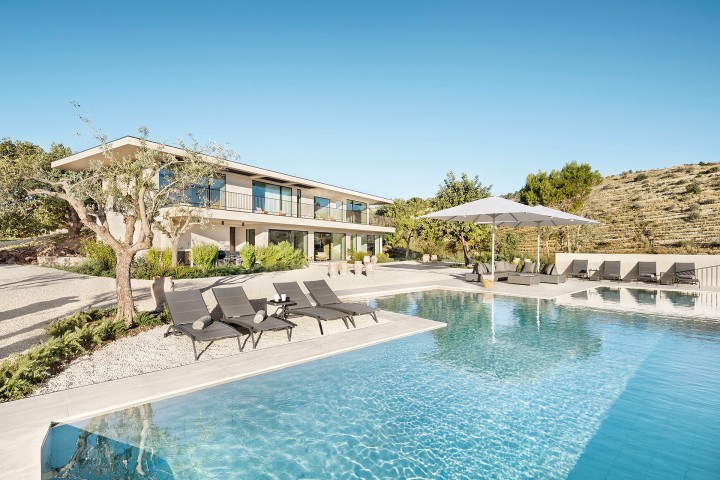 Moderne, zweistöckige Ville im Bauhausstil in mediterraner Hügellandschaft mit einem großzügigen, rechteckigen Pool gesäumt von mehreren grauen Liegen und weißen Sonnenschirmen