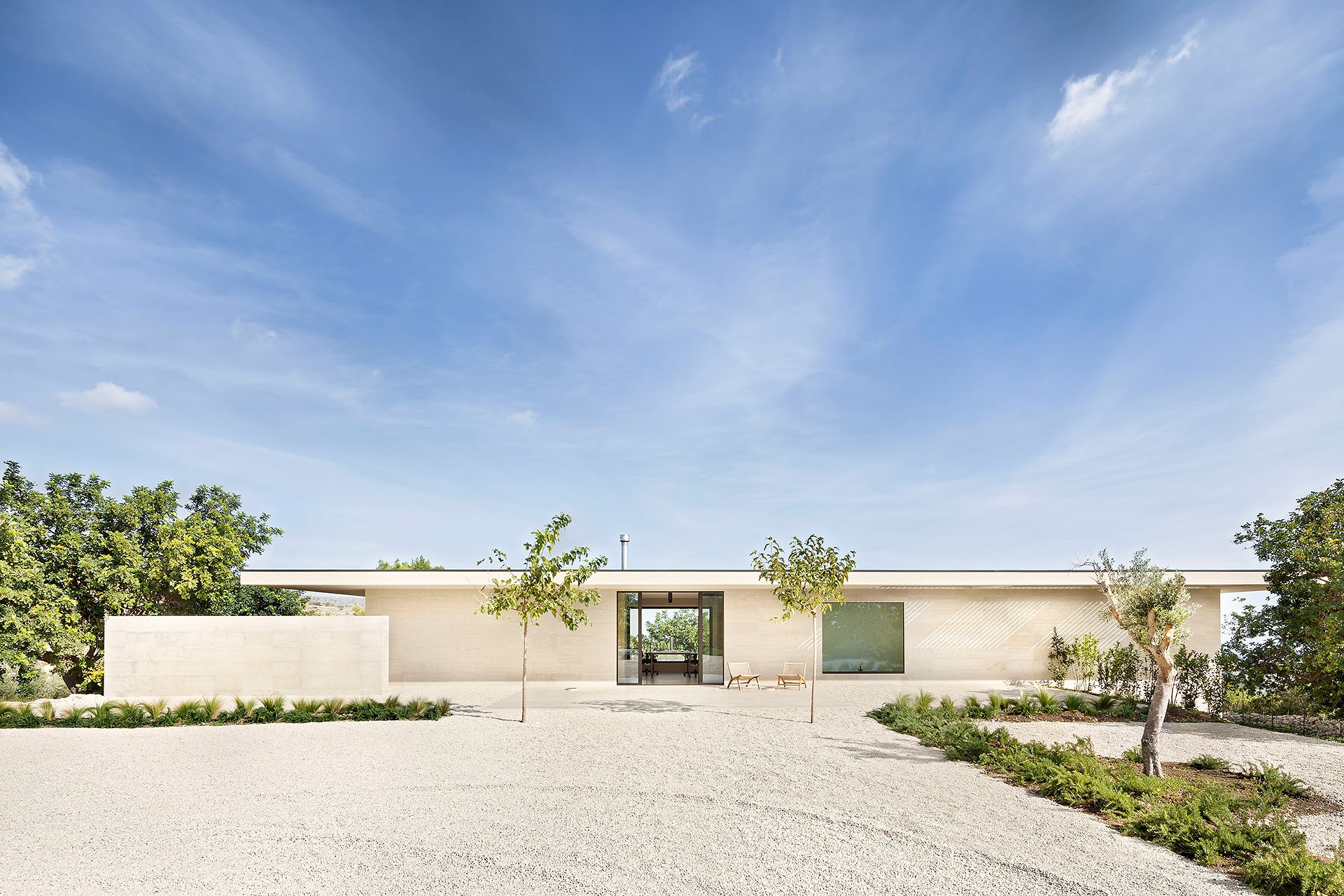 Minimalistischer Bungalow in creme mit großen, akuraten Kiesflächen und Bäumen davor im Sonnenlicht