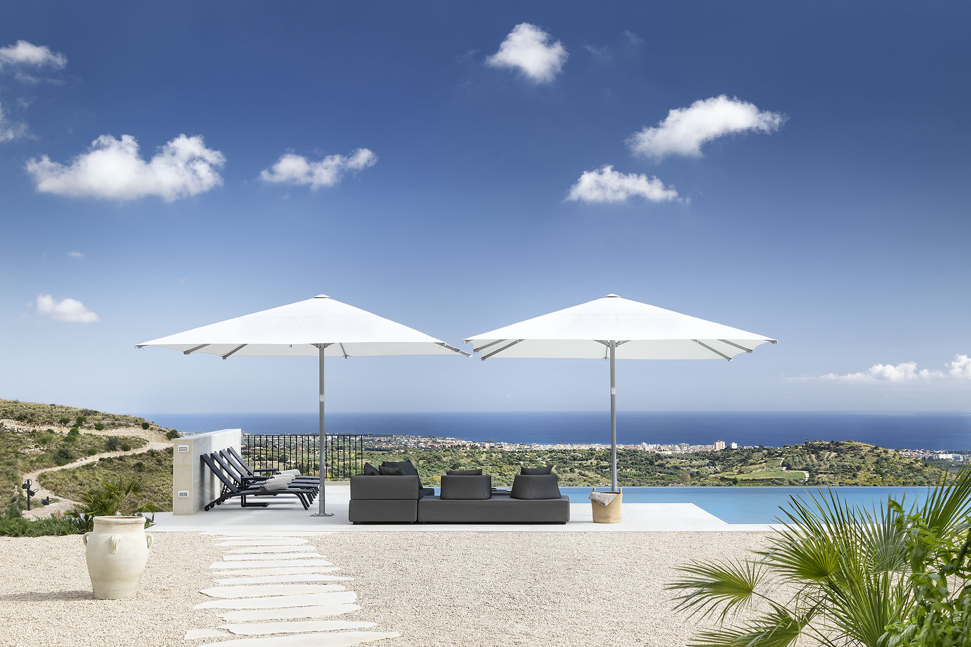 Moderne Terrasse mit Deko-Amphoren, einem rechteckigen Pool neben einer grauen Lounge-Landschaft unter zwei weißen Sonnenschirmen, im Hntergrund sonnige Hügel und das Meer