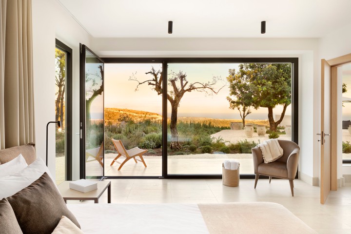 Blick aus einem modernen Wohnzimmer in hellen Naturtönen auf eine Terrasse mit Garten und Bäumen im mediterranen Stil, im Hintergrund eine Hügellandschft und dahinter das Meer im der Abendsonne