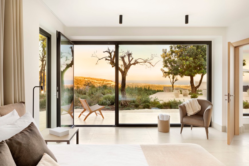Blick aus einem modernen Wohnzimmer in hellen Naturtönen auf eine Terrasse mit Garten und Bäumen im mediterranen Stil, im Hintergrund eine Hügellandschft und dahinter das Meer im der Abendsonne