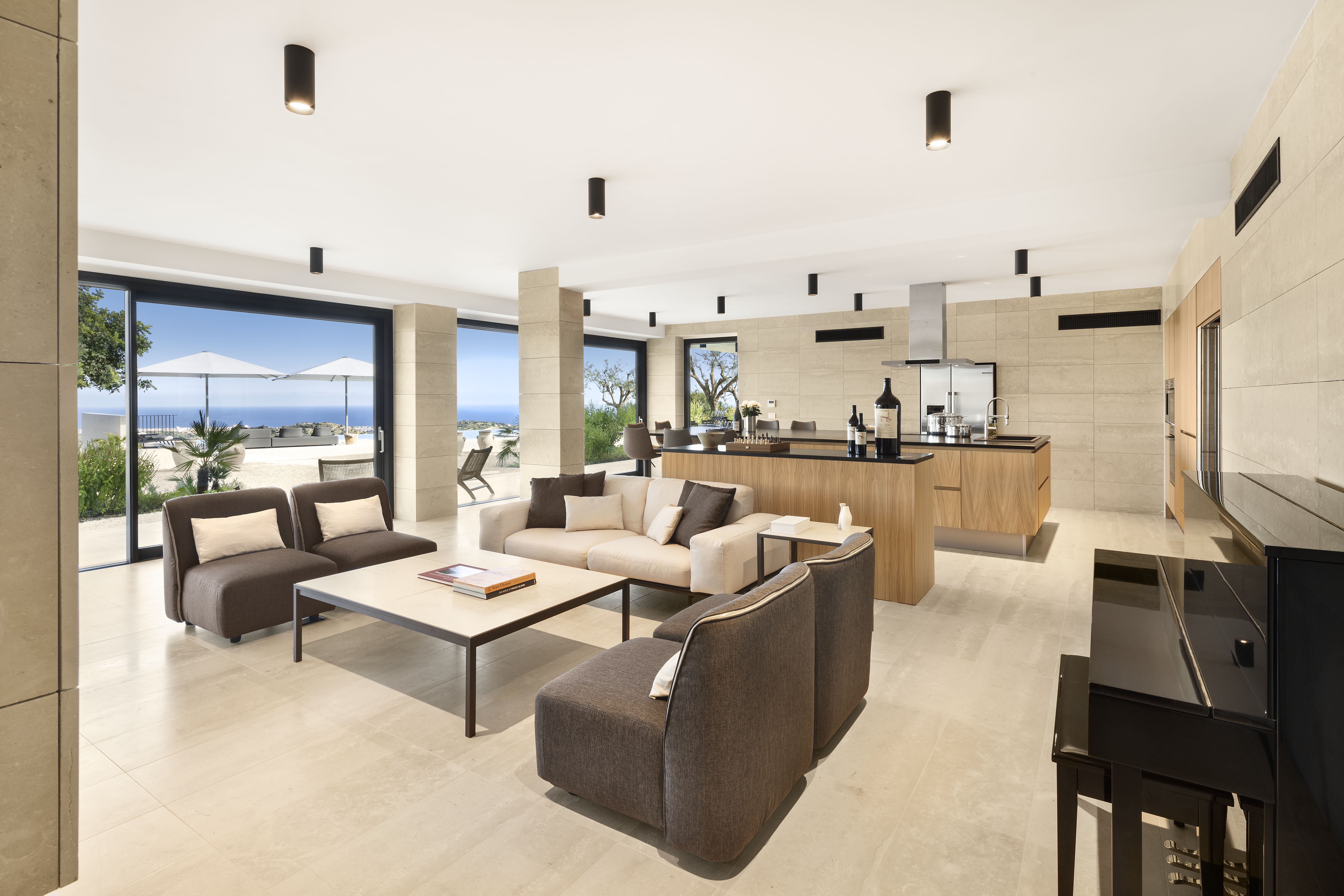 Moderne offene Küche mit Wohnbereich und Essecke in hellen Naturtönen, in der Küche stehen große Weinflaschen, im Hintergrund ist eine große Fensterfront mit Blick auf die Terrasse mit Sonnenschirm und Meer in weiter Ferne