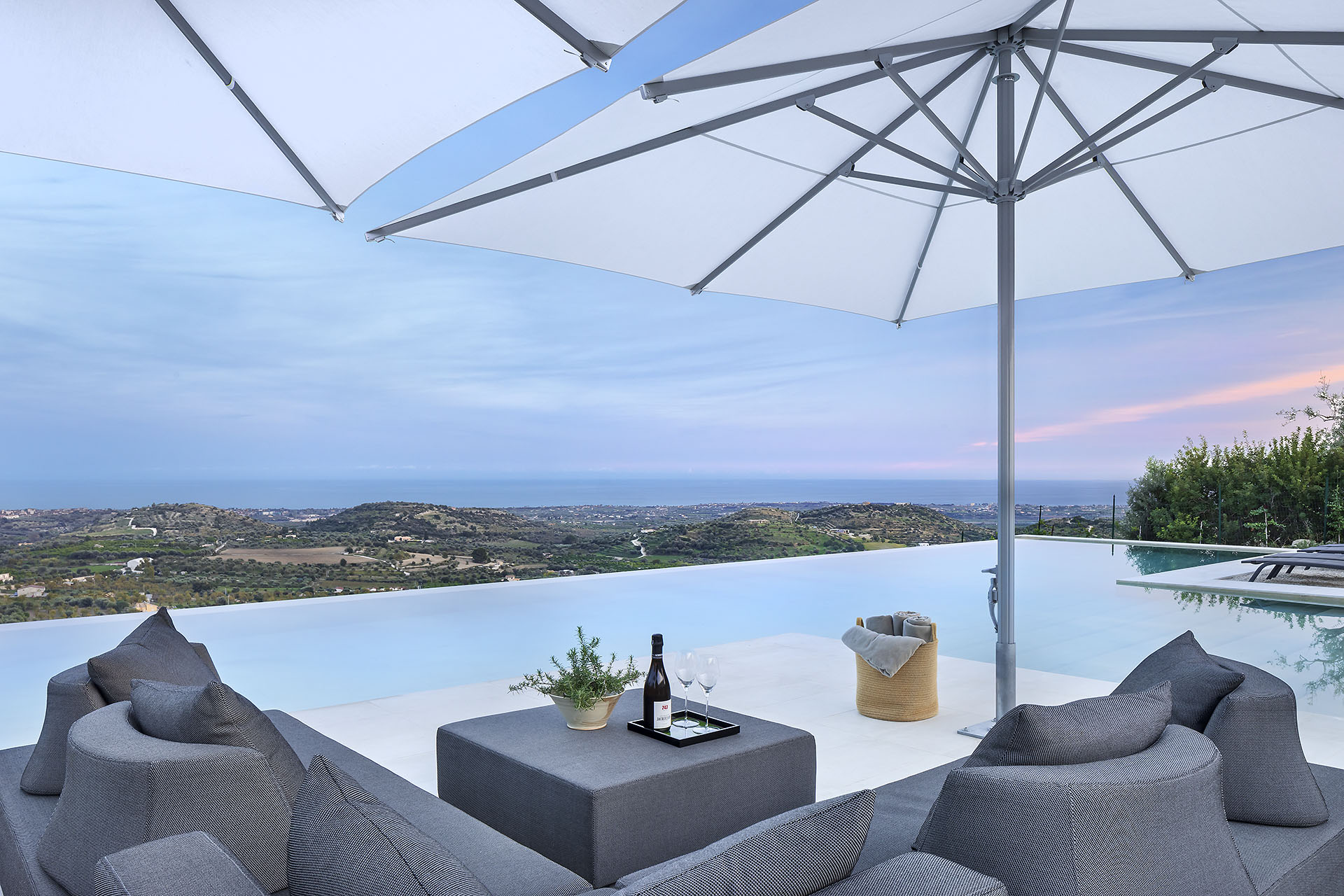 Blick von einer Terrasse mit moderner Lounge-Landschaft und Sonnenschirm über eine hügelige Insel mit Meer im Hintergrund