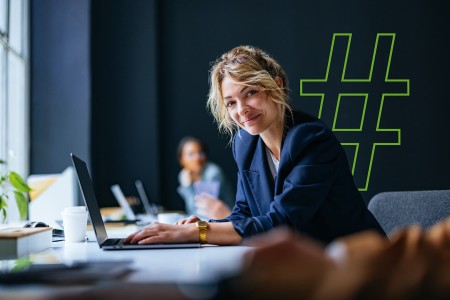 Eine Frau sitzt an einem Schreibtisch und arbeitet an einem Laptop in einem Büro. Im Hintergrund ist ein großes Hashtag-Symbol sichtbar.