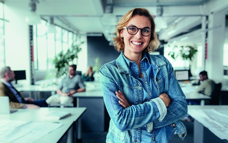 Lächelnde Geschäftsfrau mit verschränkten Armen in einem modernen Büro mit Kollegen im Hintergrund.