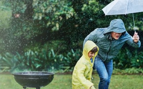 Vater und Sohn in Regenjacken und Regenschirm sprinten im Regen von einem Holzkohlegrill weg über den Rasen