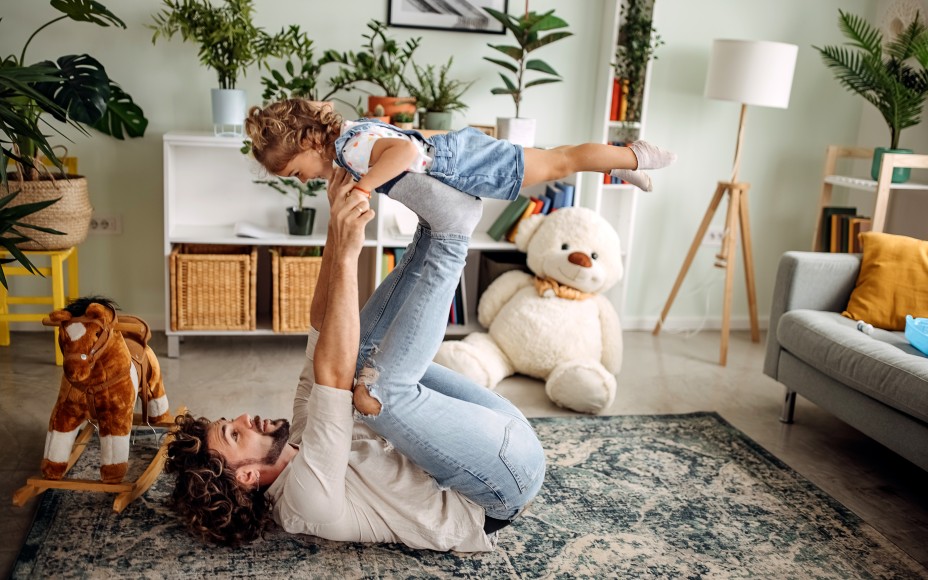 Vater liegt auf dem Rücken im Wohnzimmer und balanciert seine kleine lachende Tochter auf den Füßen, im Hintergrund eine Z8immerpflanze und eine Plüsch-Schaukelpferd