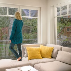 Frau mit langen blonden Haaren und grüner langer Strickjacke steht barfuß auf einem weißen Ecksofa mit gelben Kissen, das vor einer Fensterfront steht, und schaut nach draußen in ein Unwetter, beruhigt, da sie ein Elementarversicherung hat.