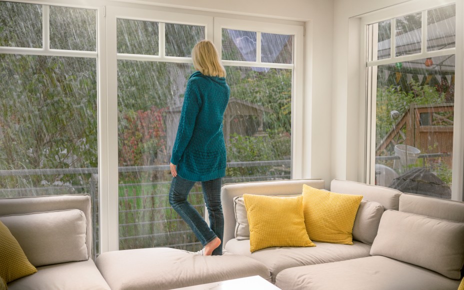 Frau mit langen blonden Haaren und grüner langer Strickjacke steht barfuß auf einem weißen Ecksofa mit gelben Kissen, das vor einer Fensterfront steht, und schaut nach draußen in ein Unwetter, beruhigt, da sie ein Elementarversicherung hat.