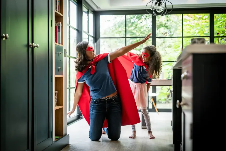 Mutter und Tochter verkleidet als Superheldinnen mit roten Umhängen und Masken in einem modernen Zuhause.