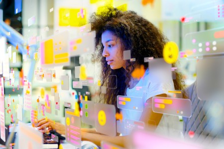 Junde Frau mit langen dunklen Locken tippt auf einer Tastatur, die zu einem Digitslrn großen Monitor gehört, auf dem viele pinke und gelbe Sticker zu sehen sind.