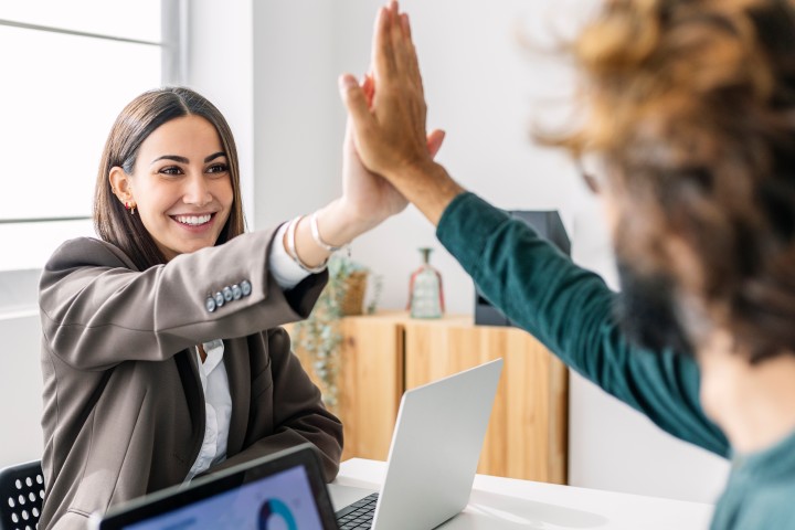 Frau mit braunem Pagenschnitt sitzt vor ihrem Laptop und gibt ihrem Kollegen lachend High Five