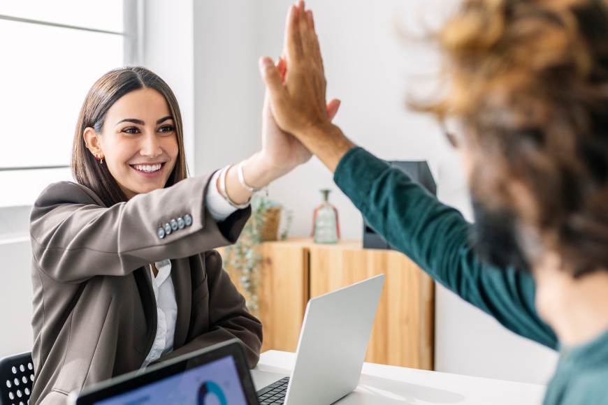 Frau mit braunem Pagenschnitt sitzt vor ihrem Laptop und gibt ihrem Kollegen lachend High Five