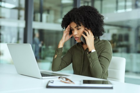Frau mit Afro-Frisur stützt den Kopf in eine Hand, mit der anderen telefoniert sie, während sie vor einem Laptop sitzt und grübelnd darauf schaut